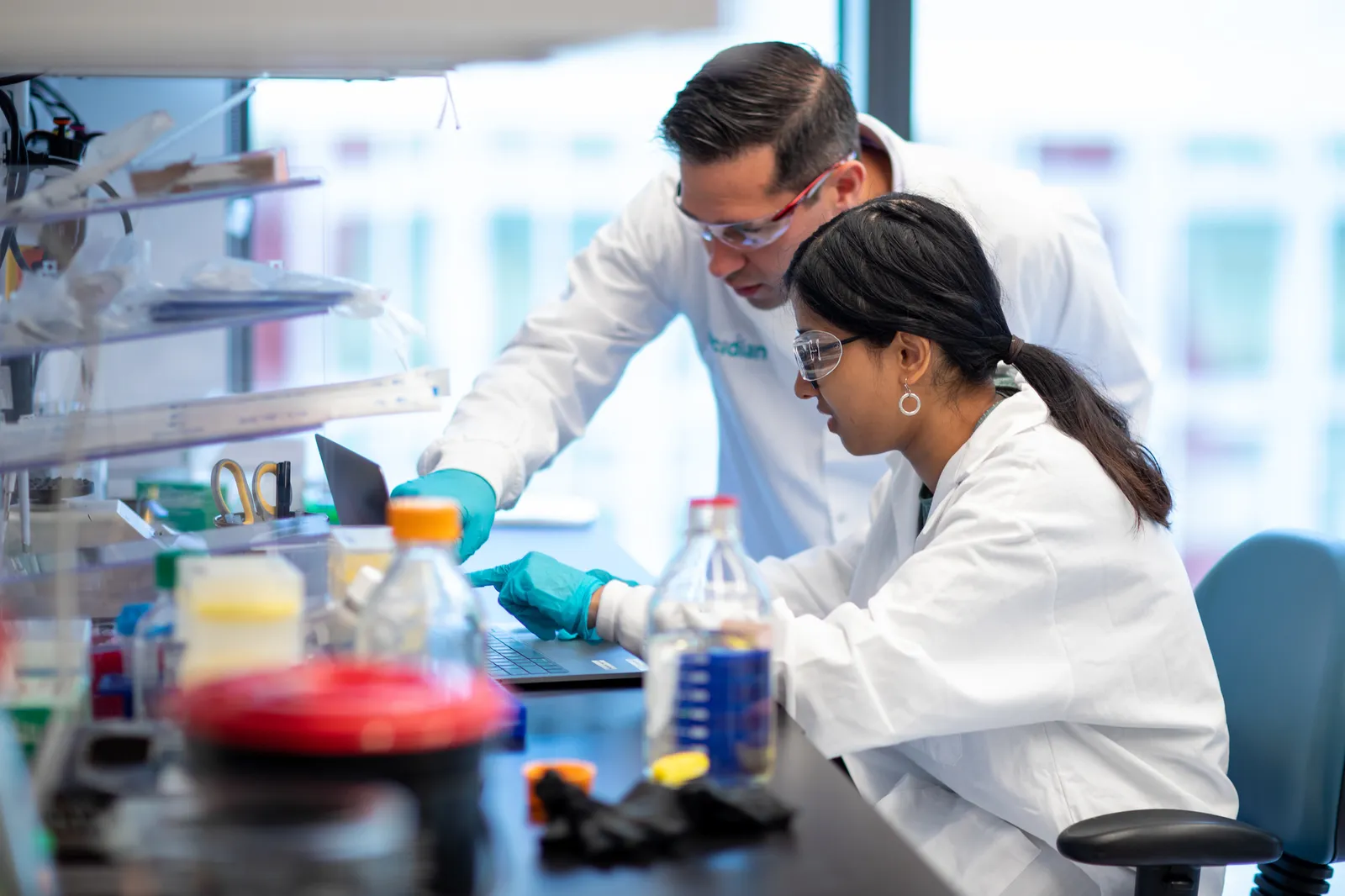 Two researchers in safety goggles and lab coats work in a lab.